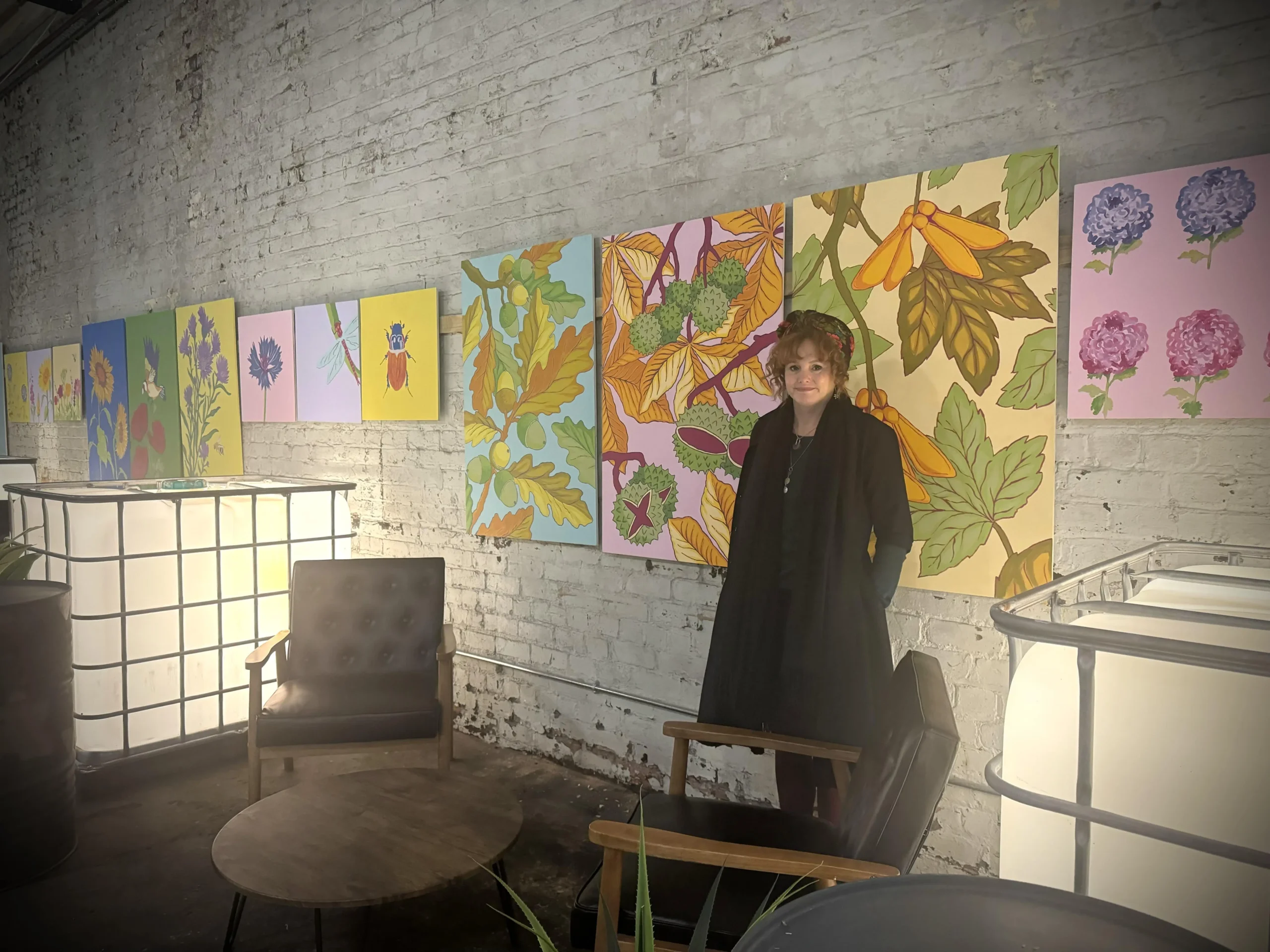 Image shows Becky stood next to three large paintings that are hanging on a wall. She is wearing a black jacket and matching headband. The paintings are of various leaves, plants, acorns and conkers. There are two arm chairs visible nearby and a small table.