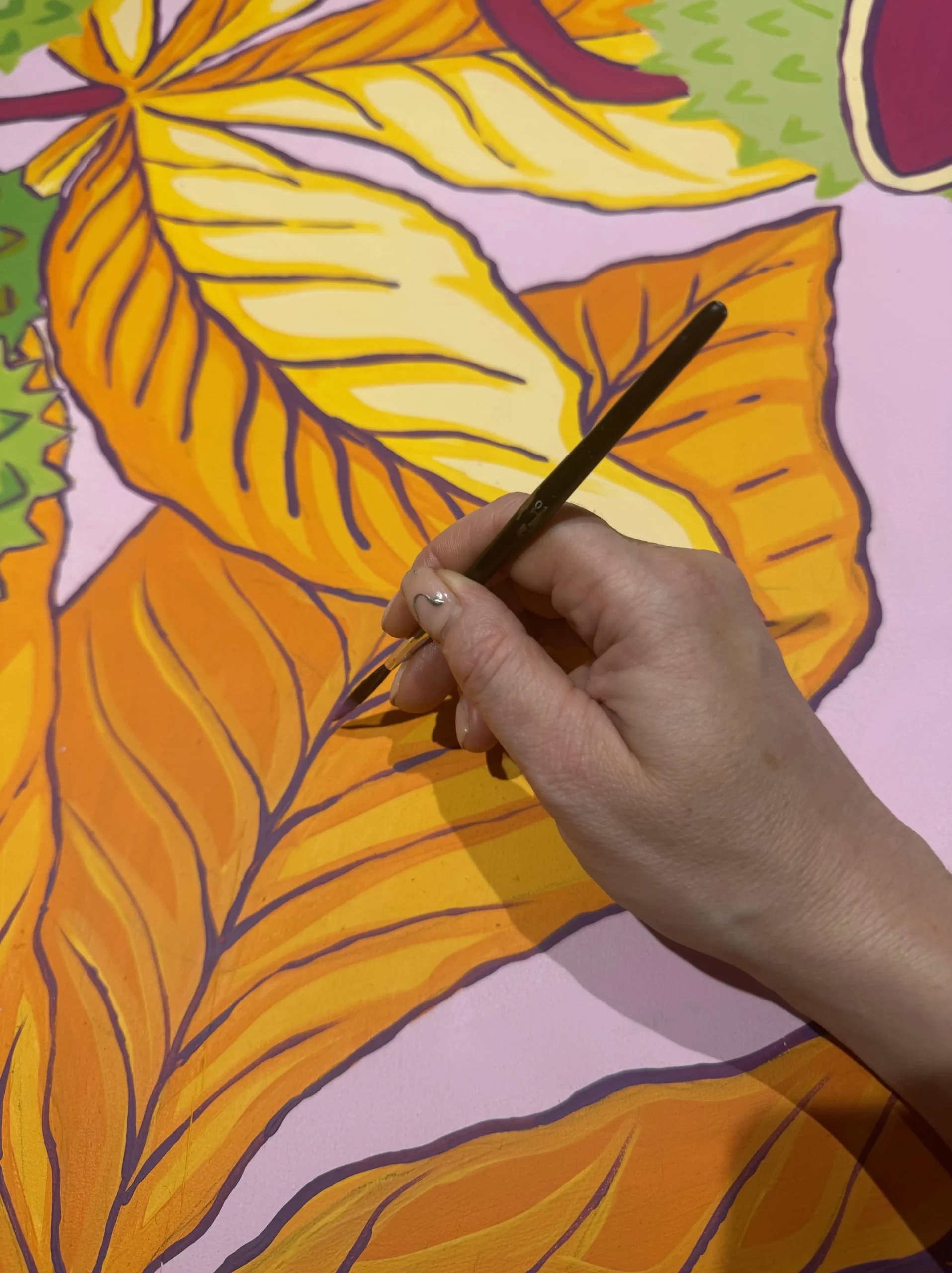 Images shows a close up of a hand, holding a paintbrush and painting on a gold and yellow leaf design.
