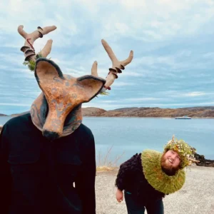 Image taken on a beach shows two people. One is wearing an animal mask with antlers and is facing the camera, the other is wearing a collar and crown made of greenary and flowers and it bend over, peering round as if to try and look at the animals face.