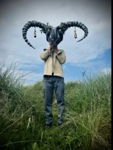 Photo is taken outdoors and shows a person standing in some long grass, wearing jeans and a beige shirt. He is holding up a large mask which is a ram with a face and large horns.